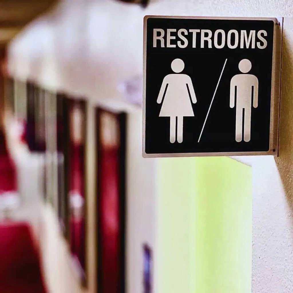 A wall-mounted restroom sign with male and female symbols and no tactile or Braille features, highlighting a common signage issue that fails to meet California Title 24 and ADA requirements for accessible restrooms.