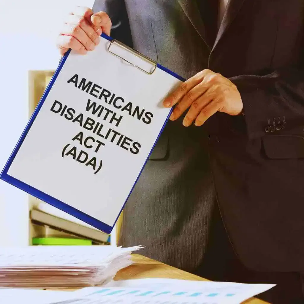 A professional in a dark suit holding a clipboard labeled “Americans with Disabilities Act (ADA)” next to a desk with building compliance paperwork symbolizing ADA and Title 24 regulatory responsibilities for California property owners.