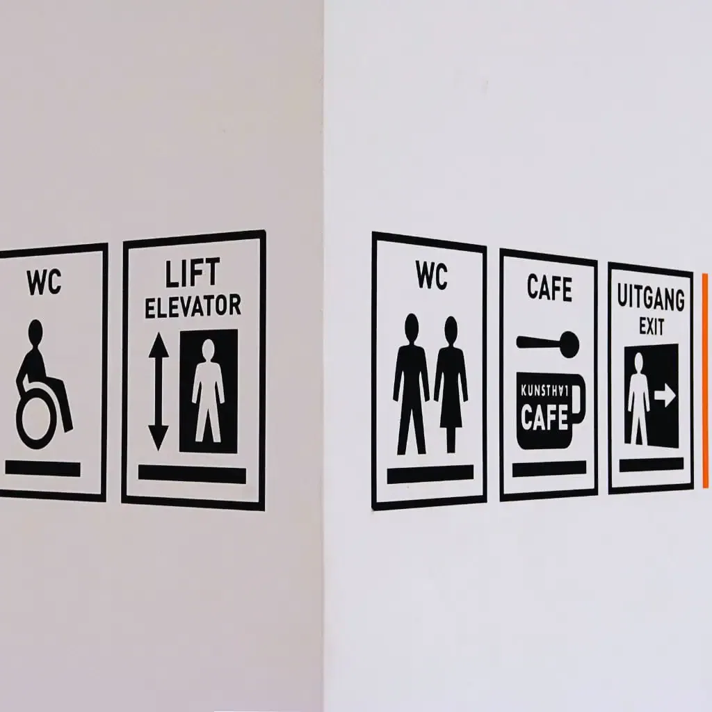 Accessible wayfinding signs on a facility wall showing restroom icons, an elevator symbol, a wheelchair-accessible restroom indicator, a café sign, and an exit symbol, representing the types of elements reviewed during a CASp inspection under California Title 24.
