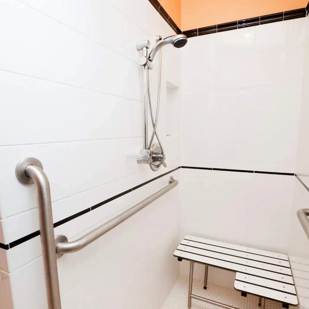 Accessible white-tiled roll-in shower with horizontal and vertical stainless steel grab bars, a handheld shower head, and a wall-mounted folding shower seat designed for ADA compliance.