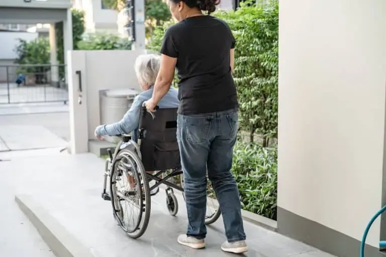 Person pushing elderly woman in wheelchair up a concrete ADA ramp near residential building