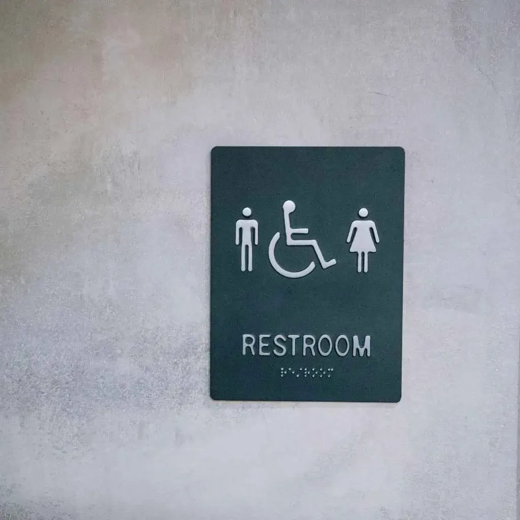 A black tactile restroom sign with male, female, and wheelchair symbols mounted on a light gray wall, representing ADA-compliant restroom signage required under California Title 24 and the 2010 ADA Standards