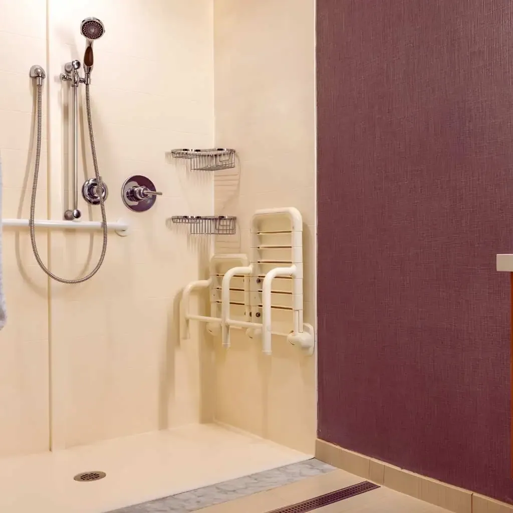Accessible roll-in shower in a California commercial building, featuring a mounted handheld showerhead, grab bars, fold-down shower seat, and wall-mounted storage baskets, meeting ADA and Title 24 standards.